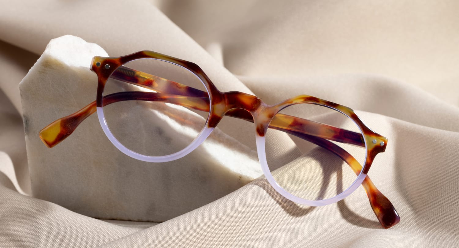 Stylish round eyeglasses with a tortoiseshell frame and translucent lower rims, displayed on a smooth beige fabric with a white marble accent, showcasing modern eyewear trends.